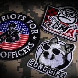 Three embroidered patches on a camo background, featuring a soldier, a monkey with a motorbike, and a corgi wearing sunglasses.