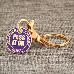 A keychain with a gold clip and a circular purple tag featuring the text "PASS IT ON" and three other words.
