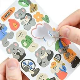 A hand holds a sheet of colourful custom stickers featuring cartoon koalas and various objects, with a small sticker partially removed.