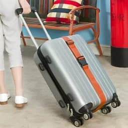A silver suitcase with an orange luggage strap being pulled along a floor, with a wooden chair in the background.