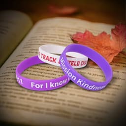 Three silicone wristbands in purple, white, and red, resting on an open book with autumn leaves. Each features a logo.