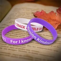 Three silicone wristbands in purple, white, and another colour with text patterns, resting on a book and autumn leaves.