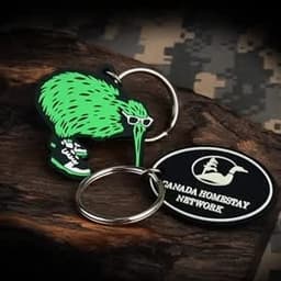 A green 2D PVC keychain shaped like a kiwi bird wearing sunglasses and sneakers, paired with a round logo keychain.