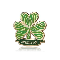 A green lapel pin shaped like a four-leaf clover, featuring a gold outline and the word "seamróg" at the bottom.
