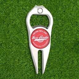 A silver divot tool with a bottle opener feature, featuring a circular logo, resting on green grass.