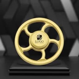 A gold metal fidget spinner with a central logo, displayed on a black pedestal against a textured background.