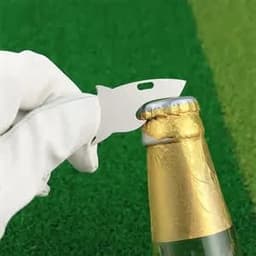 A silver divot tool shaped like a shark, positioned near a bottle cap on a green surface.
