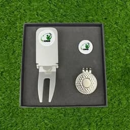 A silver golf divot tool with a pronged end, accompanied by a golf ball marker, all housed in a black box.