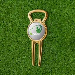 A gold metal divot tool and beer bottle opener with a circular logo, placed on green grass.