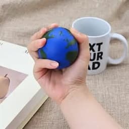 A blue and green earth-shaped stress ball being squeezed in a hand, with a mug and book in the background.