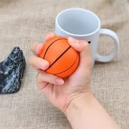 An orange basketball stress ball held in a hand, with a grey mug and rock in the background.