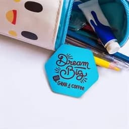 A blue hexagonal custom button featuring a design with the text "Dream Big" and a logo, resting near stationery items.