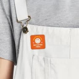 A square custom button in orange, featuring a smiley face, attached to a white overalls pocket.