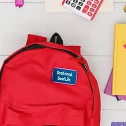 A red backpack with a blue rectangular custom button, surrounded by stationery items and colourful notebooks.