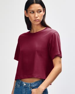 A woman with curly hair wearing a burgundy boxy t-shirt, styled with light blue jeans. The shirt has a logo.