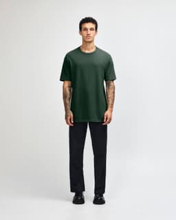 A person wearing a dark green heavyweight cotton unisex T-shirt and light blue cargo pants, standing with hands behind.