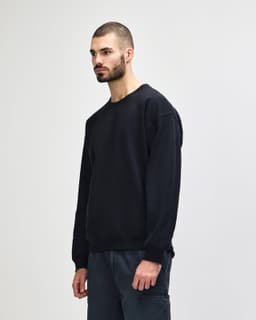 A black crewneck sweatshirt is displayed on a model standing sideways, paired with blue jeans.