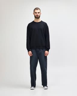 A black crewneck sweatshirt is worn by a person paired with blue jeans and white sneakers, featuring a logo.