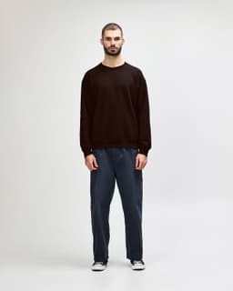 A woman wearing a dark brown Gildan Heavy Blend crewneck sweatshirt and blue jeans, standing on a neutral background.