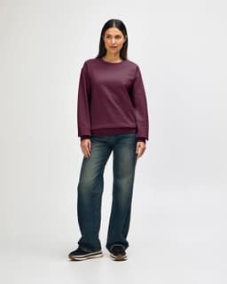 A woman stands wearing a burgundy Gildan Heavy Blend adult crewneck sweatshirt and blue jeans. The sweatshirt features a logo.