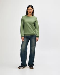 An olive green crewneck sweatshirt worn by a model, paired with black pants and sneakers. It has a logo on the sweatshirt.