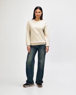 A cream-coloured crewneck sweatshirt is worn by a model paired with light blue jeans and white sneakers.