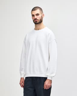 A white crewneck sweatshirt worn by a model, featuring long sleeves and a relaxed fit. It has a logo on it.
