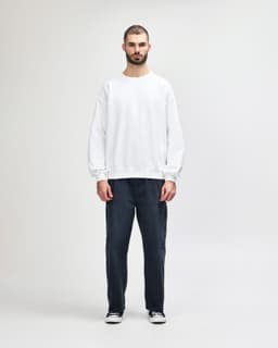 A white crewneck sweatshirt paired with black pants, worn by a model walking. The sweatshirt features a logo.