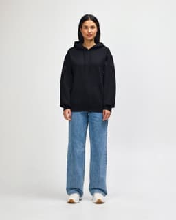 A dark grey hooded sweatshirt worn by a model with jeans and sneakers, featuring a logo on the front.