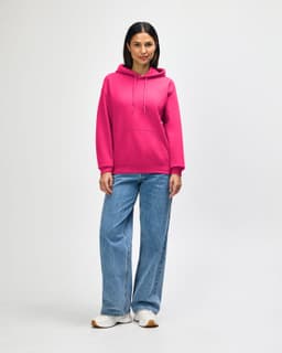 A man stands wearing a pink hooded sweatshirt with a front pocket, paired with black jeans and sneakers.