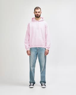 A man stands wearing a pink hooded sweatshirt and black jeans, with his hands in the pockets of the sweatshirt.