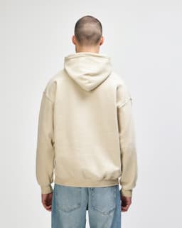 A beige hooded sweatshirt with a back view, featuring a hood and casual fit. The model wears blue jeans.