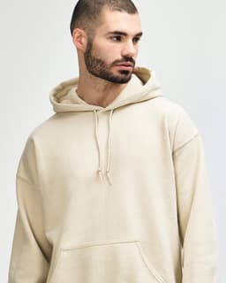 A beige hooded sweatshirt is modelled by a young woman with long hair, staring directly at the camera.