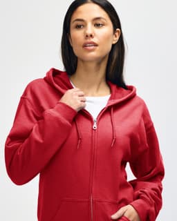 A red Gildan Heavy Blend adult full zip hooded sweatshirt on a model adjusting the hood. The sweatshirt features a logo.