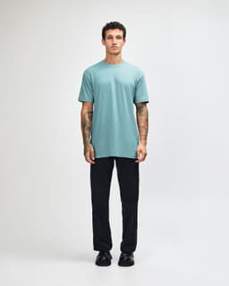 A unisex short sleeve t-shirt in a teal colour, paired with black cargo pants and casual shoes. The model has a relaxed pose.