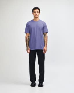 A unisex short sleeve t-shirt in purple, worn by a model with black pants and sneakers, featuring a logo.