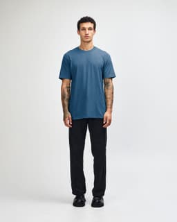 A unisex CVC short sleeve t-shirt in a blue shade, paired with black cargo pants. The model stands casually.
