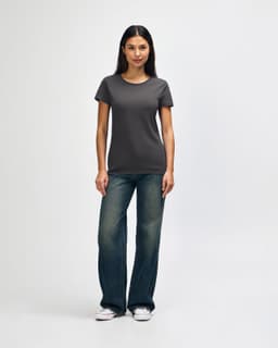 A woman wearing a dark grey Gildan Heavy Cotton ladies' T-shirt, paired with blue jeans and white sneakers.