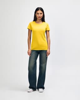 A woman wearing a yellow Gildan Heavy Cotton ladies' t-shirt paired with blue jeans and white sneakers.