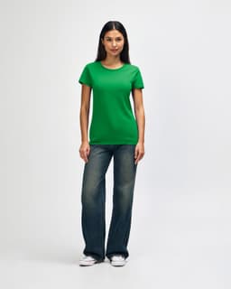 A woman stands wearing a green Gildan Heavy Cotton Ladies' T-Shirt and blue jeans, with a casual pose and white shoes.