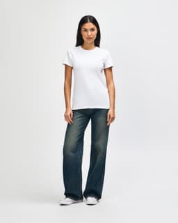 A woman stands in a white Gildan Heavy Cotton Ladies' T-Shirt paired with blue jeans and white sneakers.