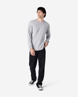 A model wearing a grey long sleeve t-shirt paired with black trousers and sneakers, standing against a plain background.