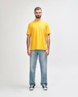 A man stands wearing a bright yellow Gildan Softstyle adult T-shirt paired with grey jeans. The shirt has a logo.