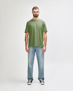A man stands wearing an olive green Gildan Softstyle adult T-shirt paired with light blue jeans.