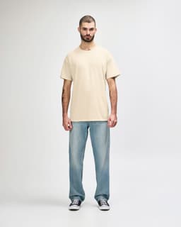 A model stands wearing a cream Gildan Softstyle adult T-shirt paired with light blue jeans and white sneakers.
