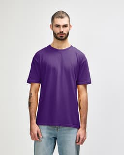 A man stands wearing a purple Gildan Softstyle adult T-shirt, paired with light blue jeans. The shirt has a logo.