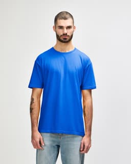 A man models a blue Gildan Softstyle adult t-shirt, paired with light denim jeans. The shirt has a logo.