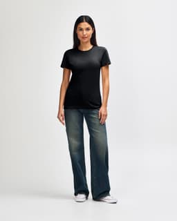 A model wears a black Gildan Softstyle ladies' T-shirt paired with blue jeans and white sneakers, showcasing a casual look.