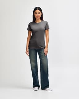A woman stands wearing a soft-style ladies' t-shirt in grey, paired with blue jeans and white sneakers.