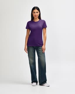 A woman is wearing a purple Gildan Softstyle ladies' T-shirt paired with blue jeans and white sneakers.